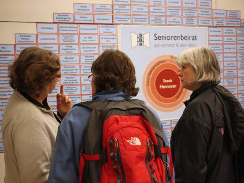 Drei Damen diskutieren vor einer Wand mit der Übersicht ehrenamtlicher Tätigkeiten von Seniorinnen und Senioren in Hannover